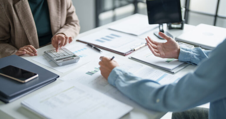 Two people discussing financial documents at a desk with a calculator and laptop.