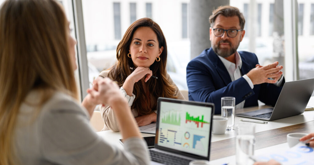 Business meeting with three people discussing financial data on laptops and tablets.