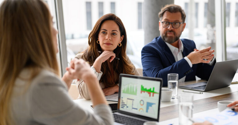 Business meeting with three people discussing financial data on laptops and tablets.